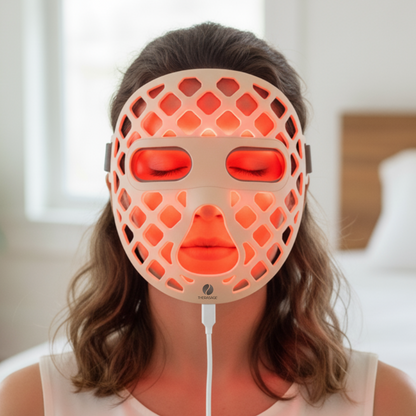 A woman wears the glowing red TheraGlow Mask2Go+™ LED Light Therapy Face Mask, facing forward indoors.