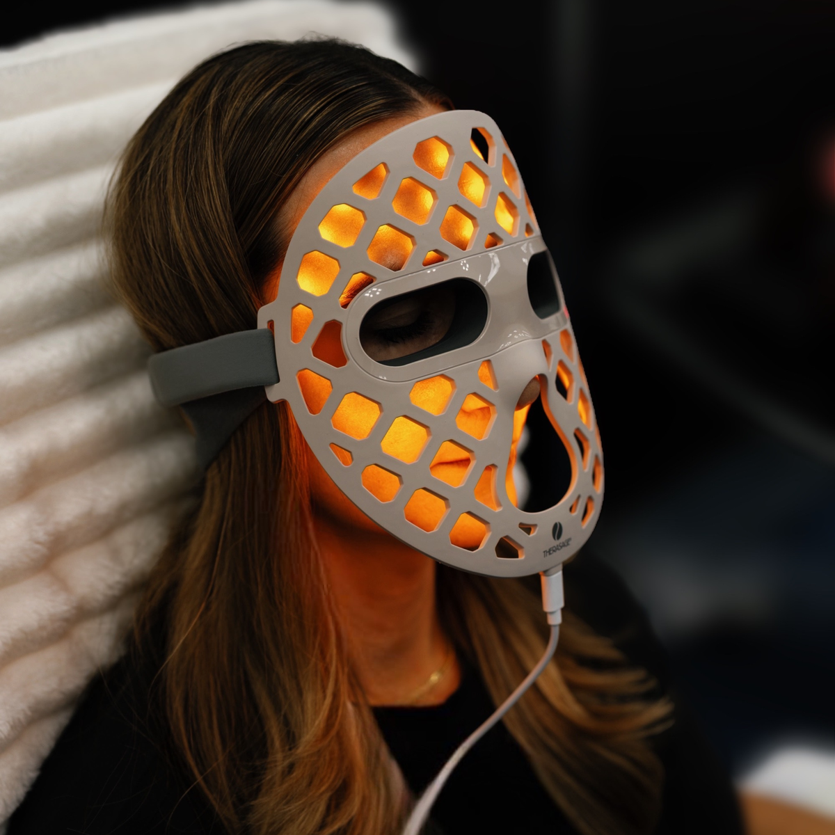 A woman reclines wearing the amber-lit TheraGlow Mask2Go+™ LED Light Therapy Face Mask, viewed from the side.