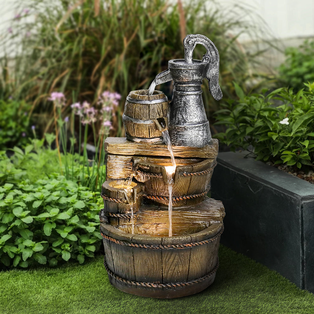 Resin Broken Barrel Rustic Outdoor Fountain 25 Inch LED stands outdoors on grass near a garden bed.