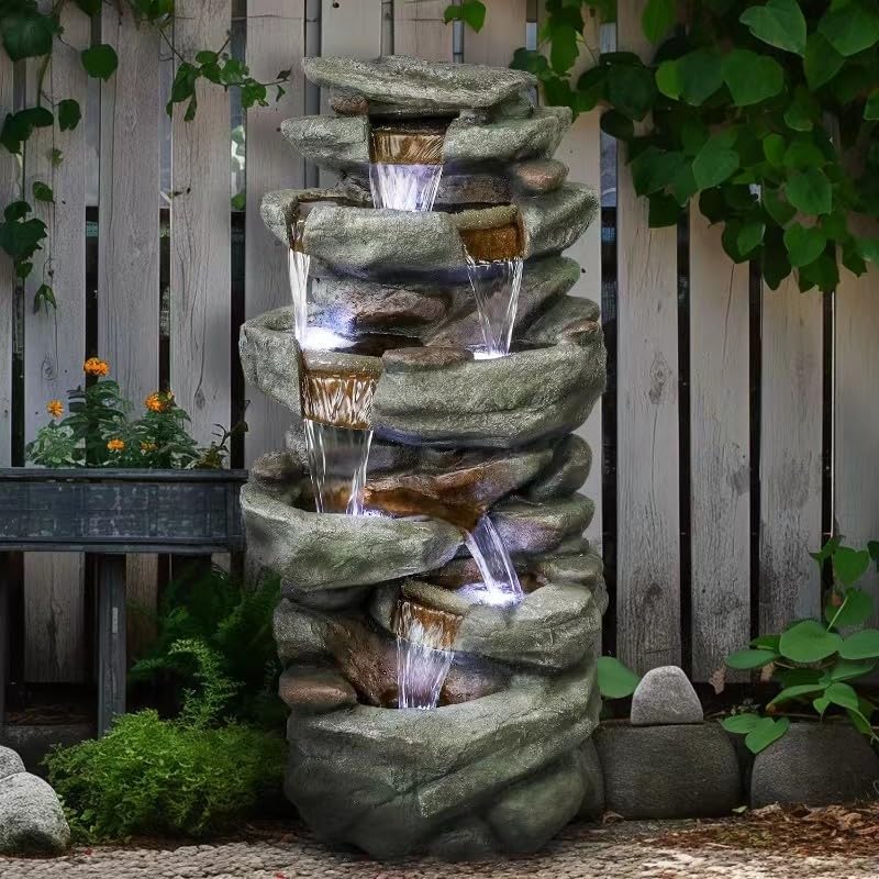 Resin Overhanging Ledge Rock Waterfall Fountain Outdoor with cascading water beside a garden bench and leafy fence.