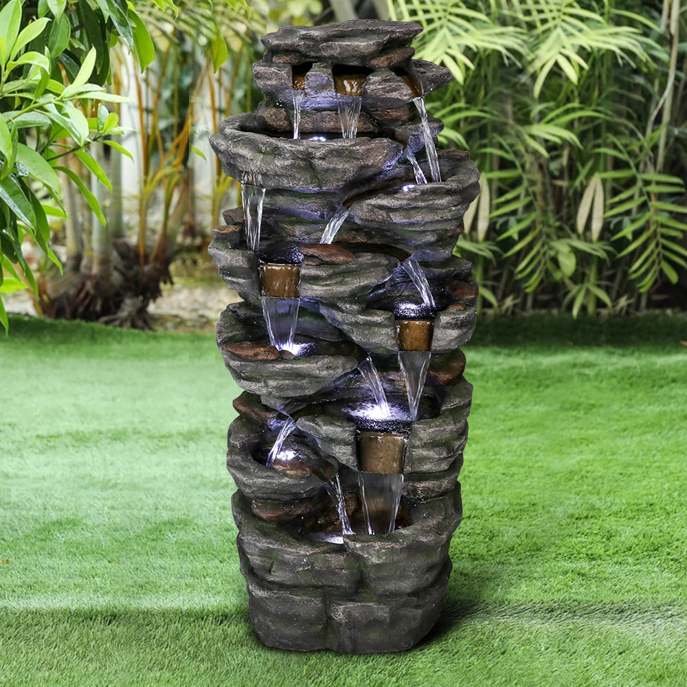 Resin Jagged Shale Outdoor Waterfall Fountain 13 Streams cascades on a green lawn amid leafy garden plants.