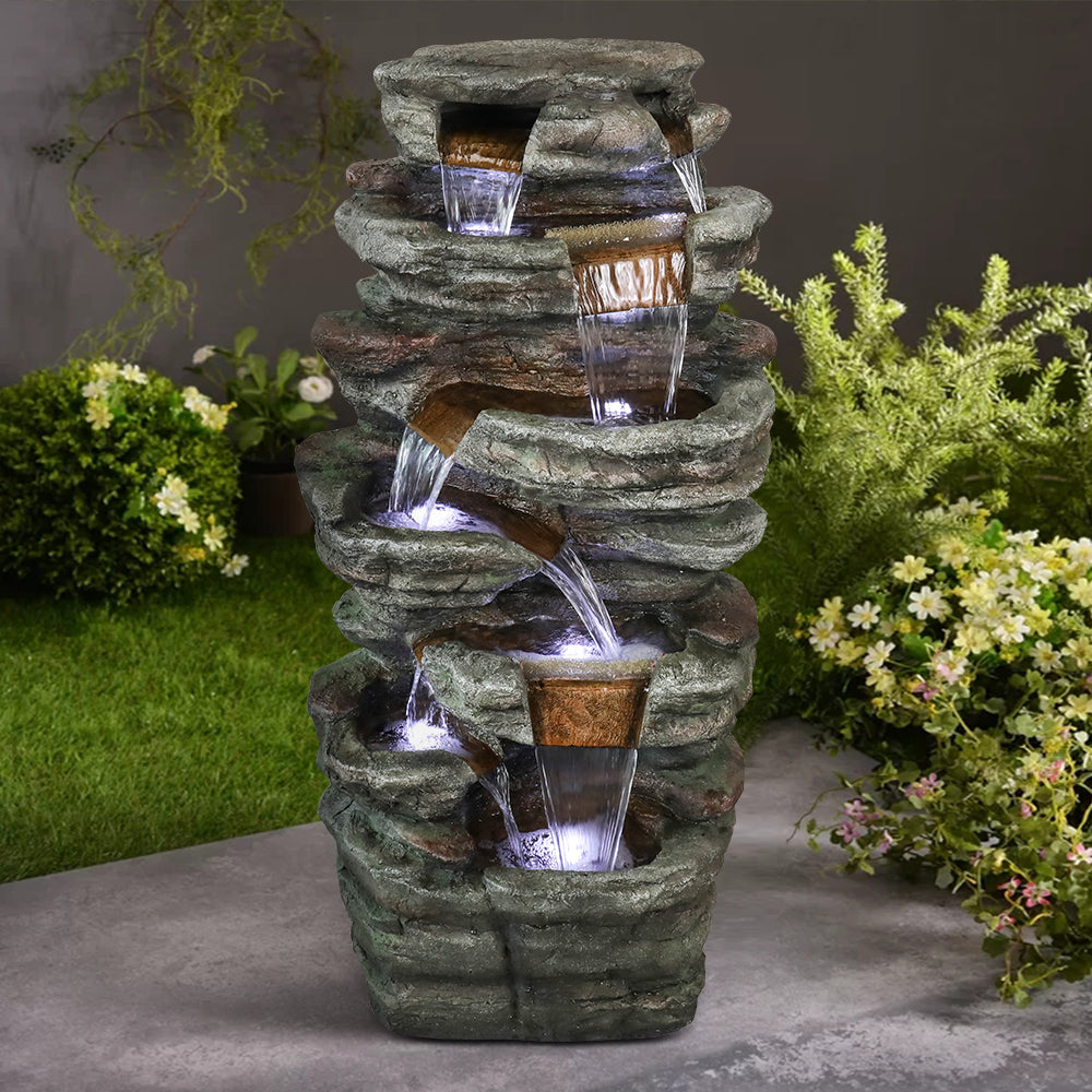 Resin Serpentine Slate Column Cascading Outdoor Fountain on a patio, framed by flowering shrubs and lawn.