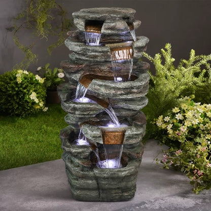 Resin Serpentine Slate Column Cascading Outdoor Fountain on a patio, framed by flowering shrubs and lawn.