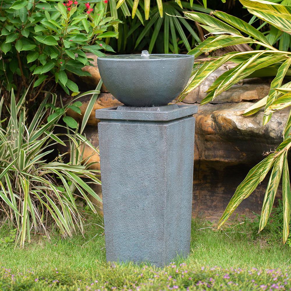 Resin Zen Bowl Pedestal Outdoor Fountain 35 Inch bubbling in a lush garden among rocks and leafy plants.