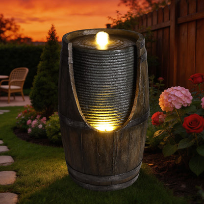 Resin Rustic Barrel Outdoor Water Fountain 24 Inch LED glowing in a garden at sunset, surrounded by flowers.