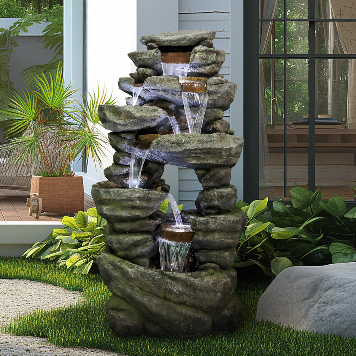 Resin Rock Waterfall Fountain Large 60 Inch 7 Tier flows beside a patio window, potted palm, and garden plants.