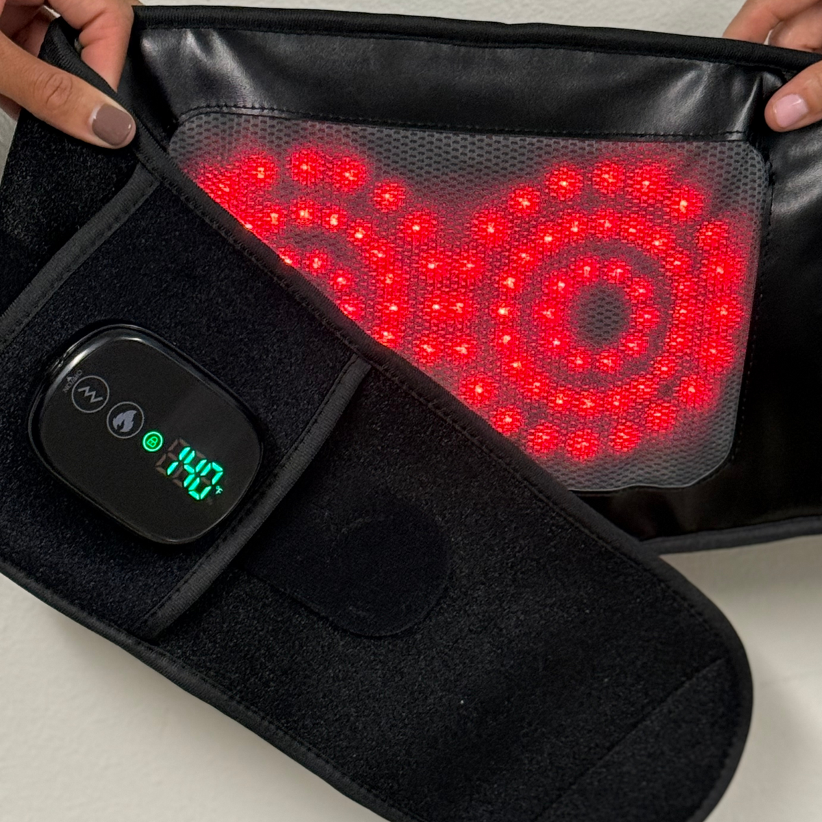 Hands hold open the TheraBelt Red Light Therapy Recovery Belt, revealing its glowing red panel and green-lit controller.