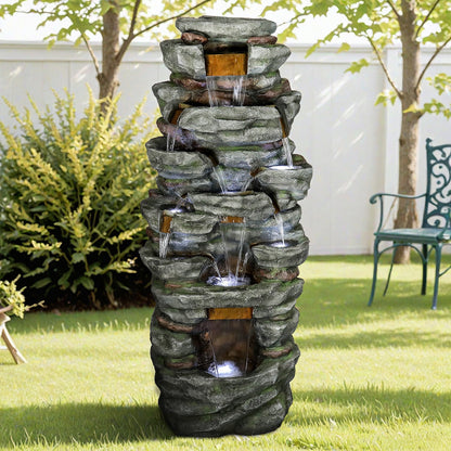 Resin Cliffside Rock Large Outdoor Water Fountain on a lawn with a green bench, fence, and trees.
