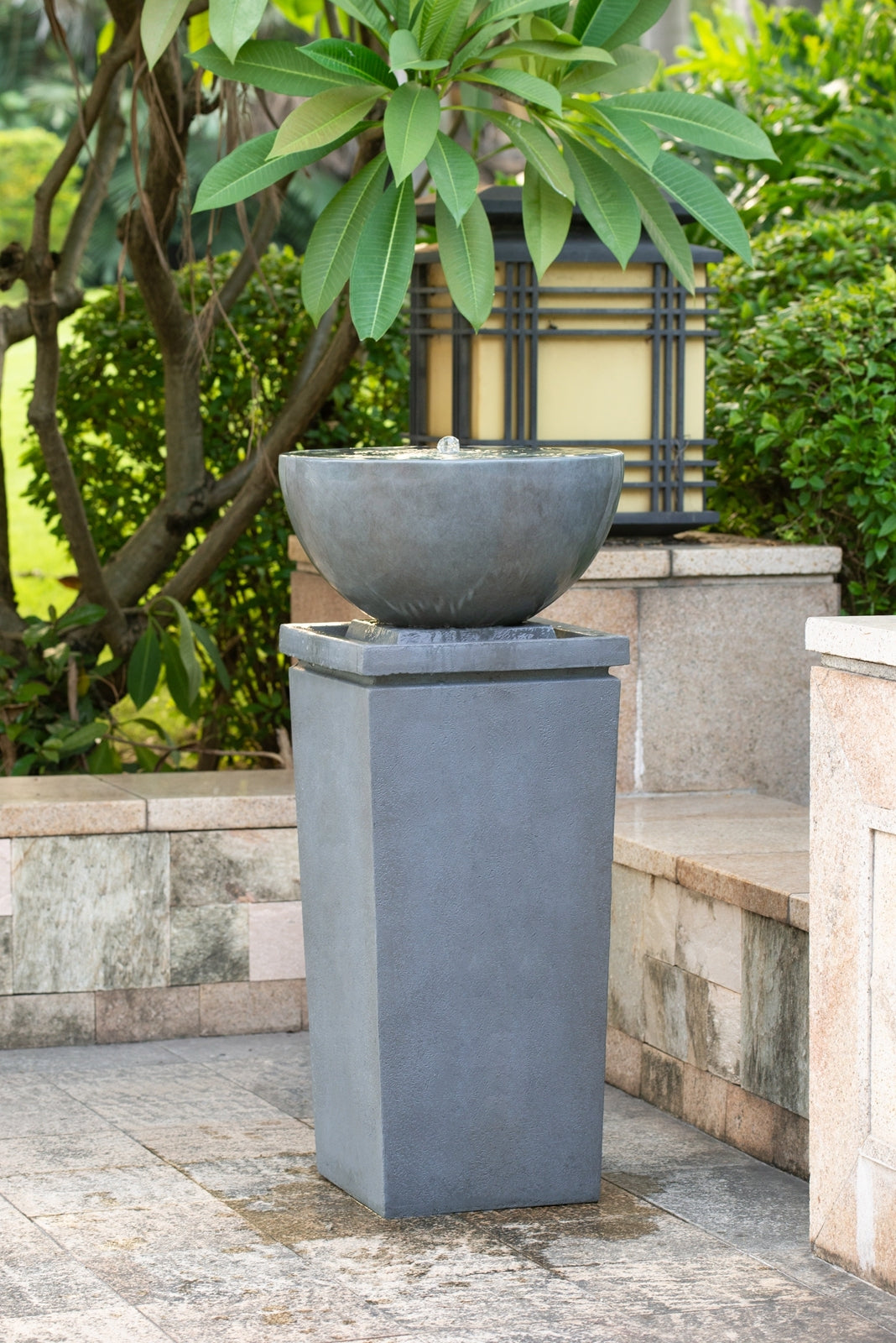 Resin Zen Bowl Pedestal Outdoor Fountain 44 Inch stands on a stone patio among greenery and a garden lantern.