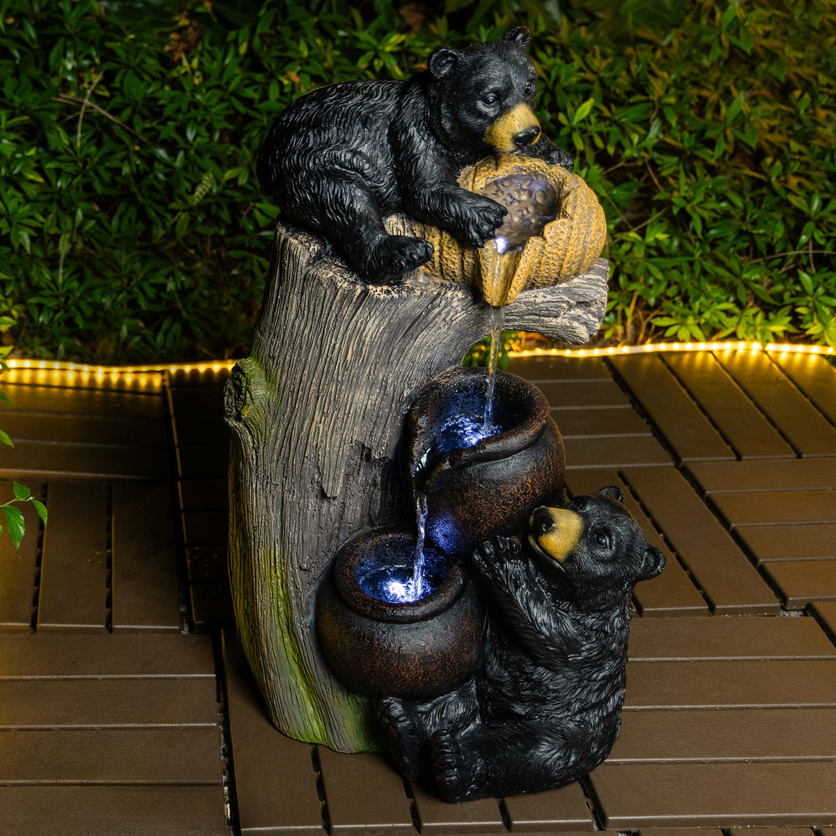 Resin Black Bear Outdoor Water Fountain with blue-lit bowls on a brown deck by dense greenery.