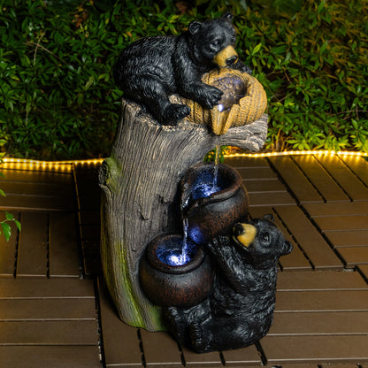 Resin Black Bear Outdoor Water Fountain with blue-lit bowls on a brown deck by dense greenery.