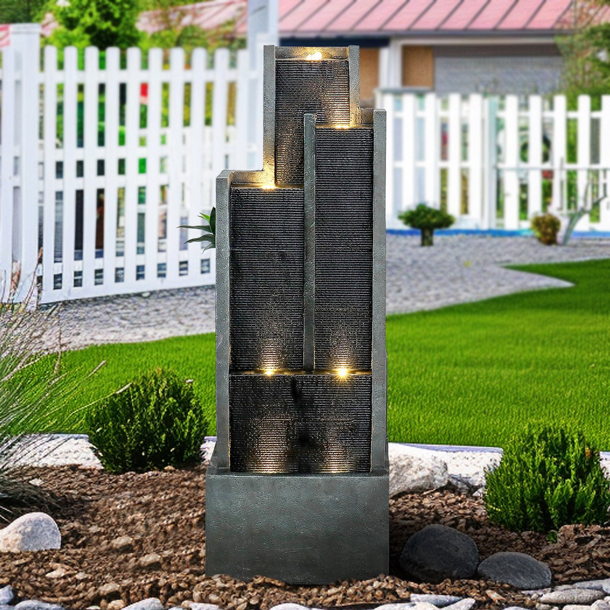 Resin Ribbed Slate Outdoor Water Fountain Quiet 50 in stands among lawn rocks and shrubs beside a white garden fence.