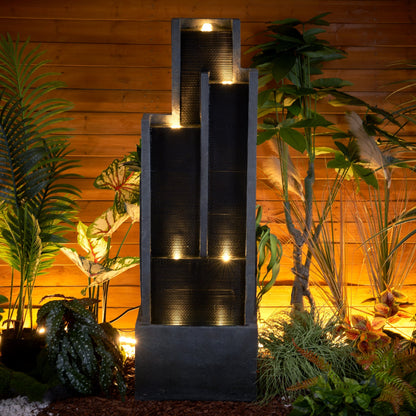 Resin Ribbed Slate Outdoor Water Fountain Quiet 50 in glows at night in a planted garden beside a wood fence.