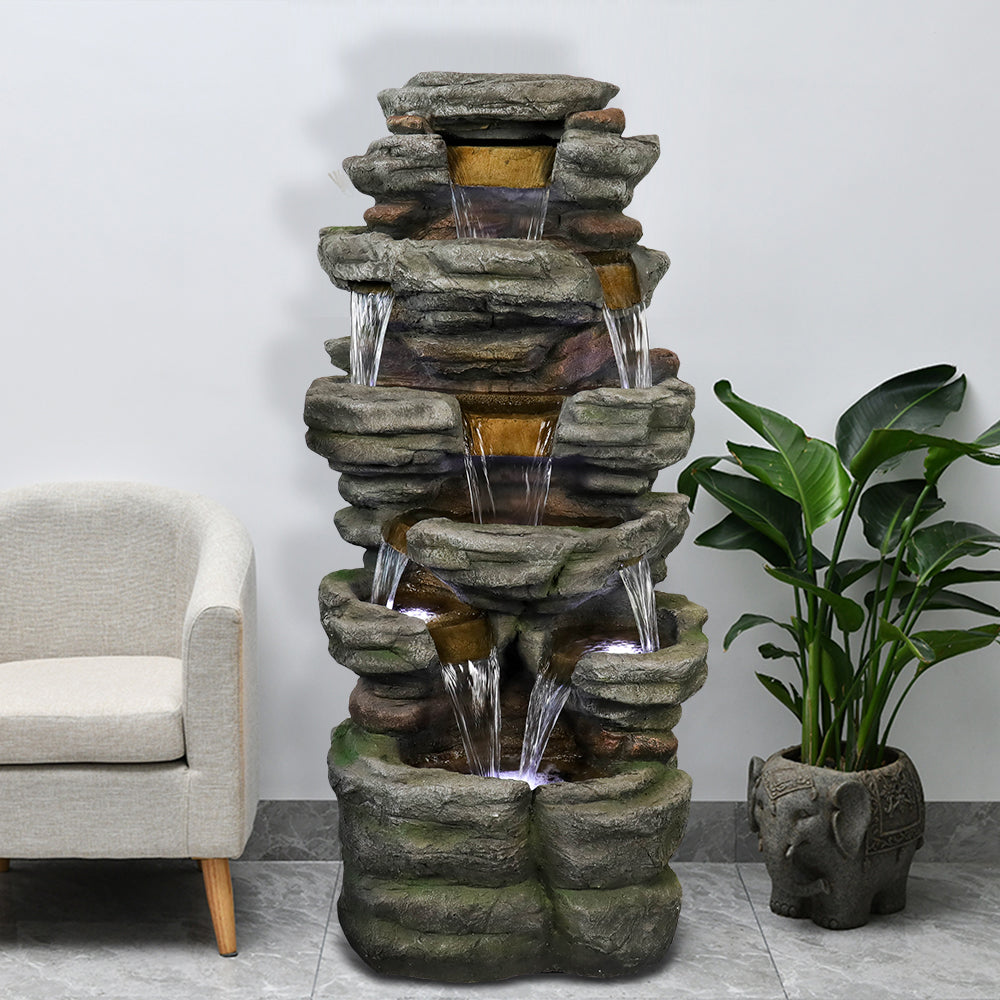 Resin Stepped Shale Outdoor Rock Fountain with cascading water indoors, beside a white chair and leafy plant.