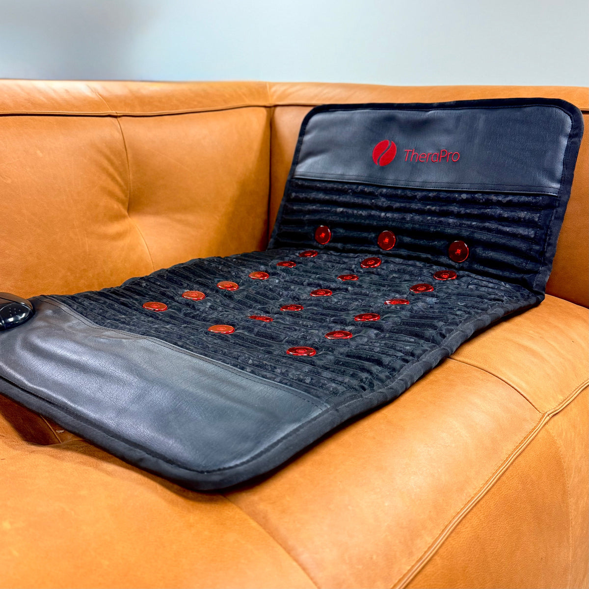 TheraPro™ PEMF pad unfolded across an orange leather couch, with red circular elements and a controller at the side.