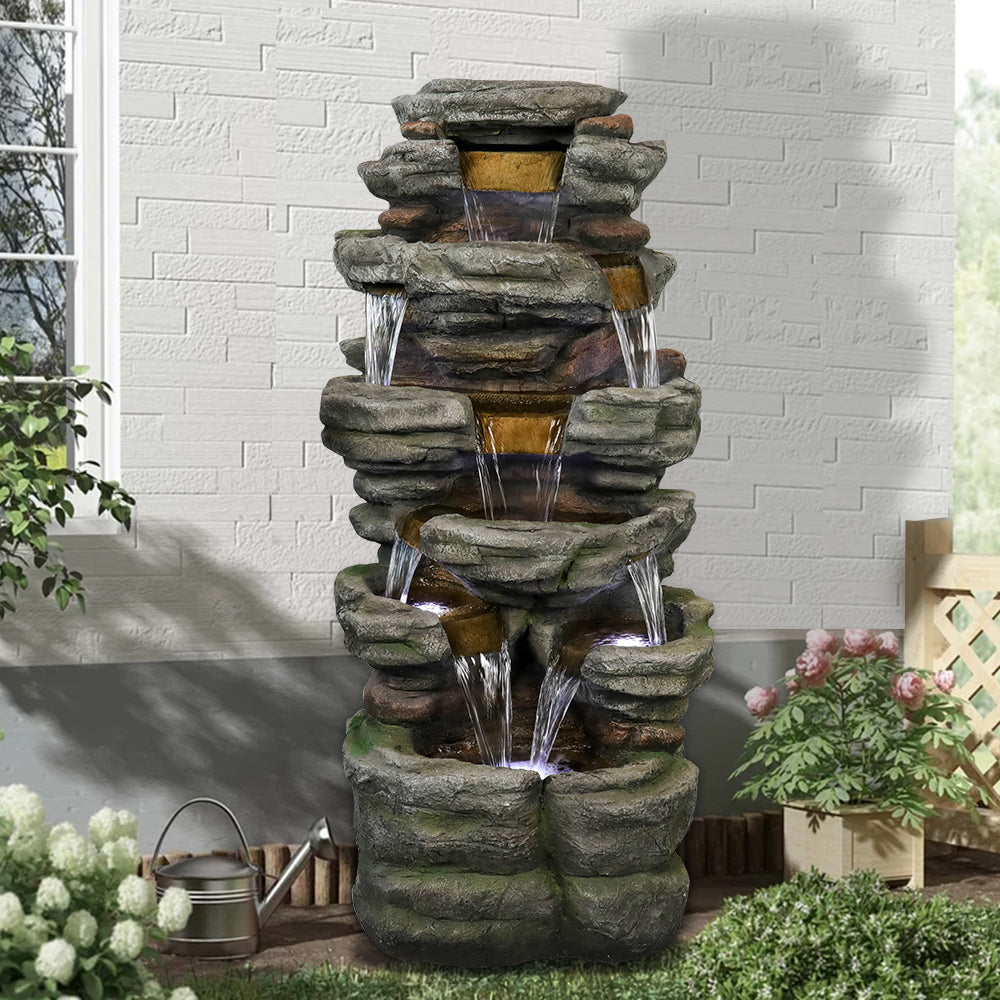 Resin Stepped Shale Outdoor Rock Fountain pours into tiered basins beside a garden wall and potted flowers.