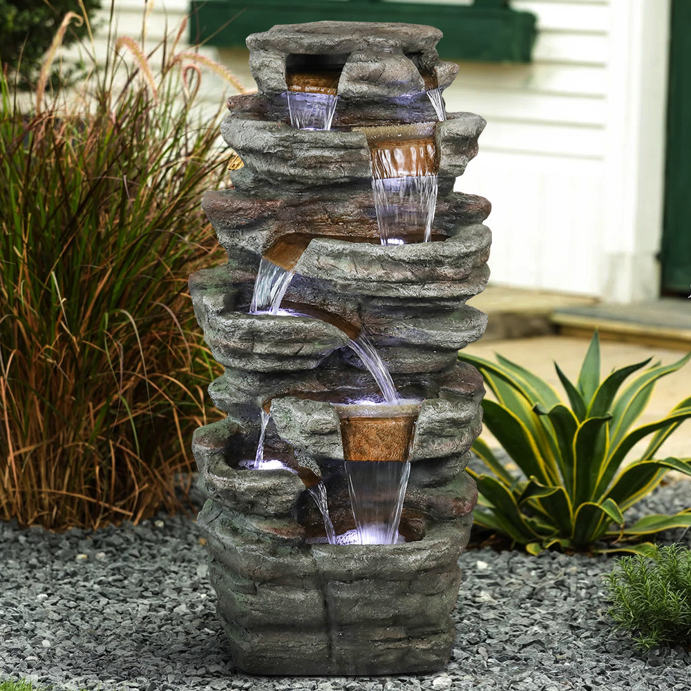Resin Serpentine Slate Column Cascading Outdoor Fountain surrounded by gravel and garden plants beside a house.