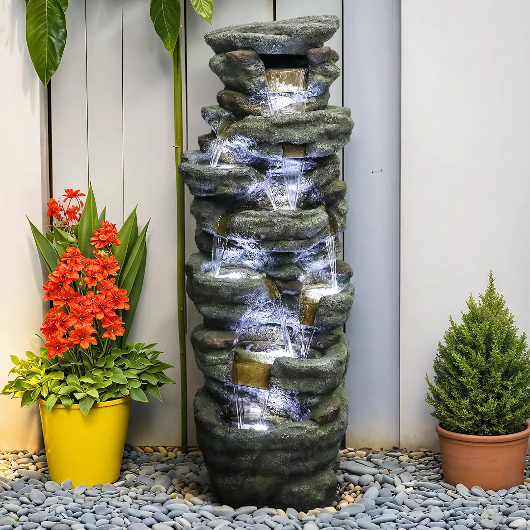 Resin River Rock Outdoor Fountain Pillar 51 Inch 52 lb glows with lights as water cascades over tiers between potted plants.