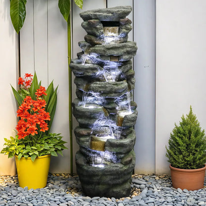 Resin River Rock Outdoor Fountain Pillar 51 Inch 52 lb glows with lights as water cascades over tiers between potted plants.