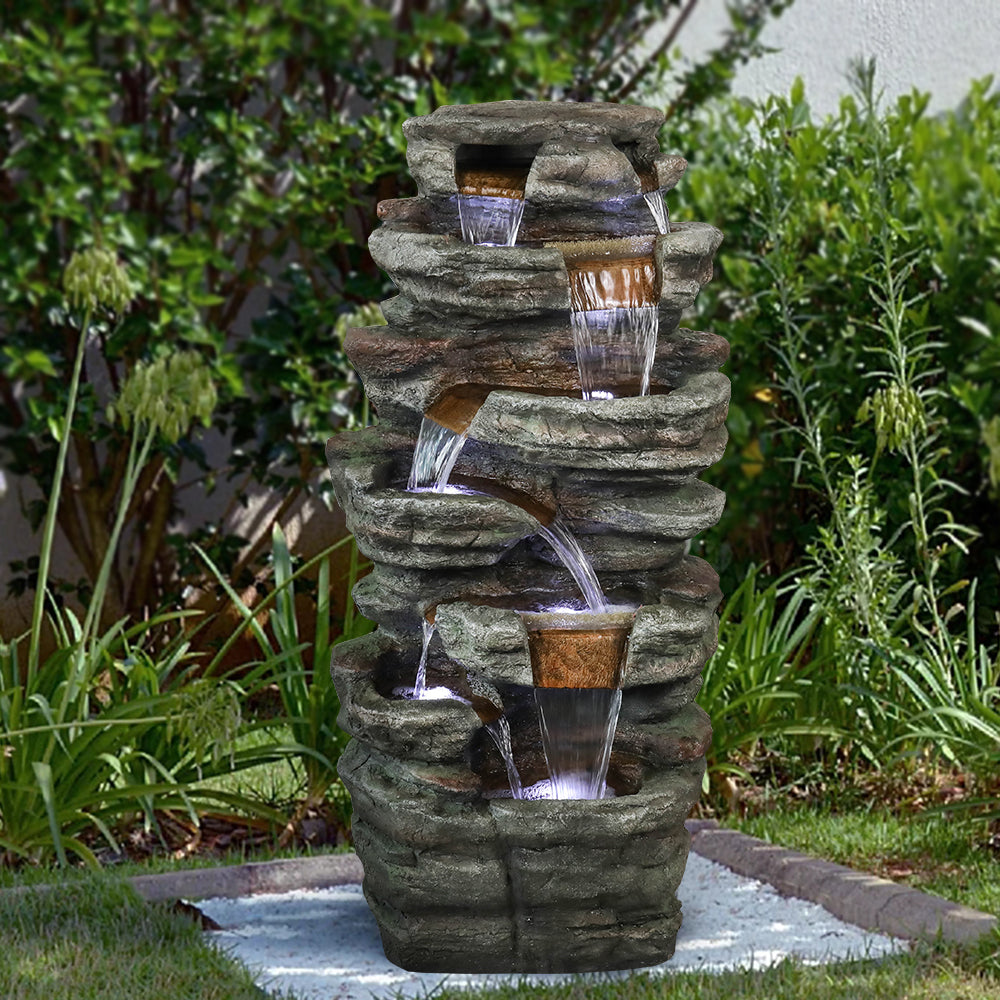Resin Serpentine Slate Column Cascading Outdoor Fountain centered among dense green garden plants.