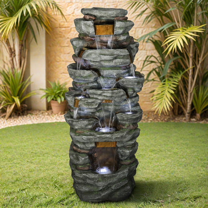Resin Cliffside Rock Large Outdoor Water Fountain with cascading water, set on a lawn among palms and a stone wall.