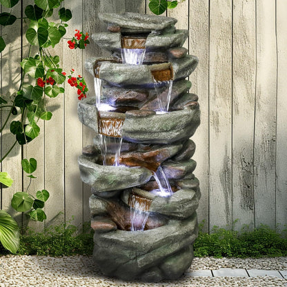 Resin Overhanging Ledge Rock Waterfall Fountain Outdoor shown flowing in front of a pale fence and climbing greenery.