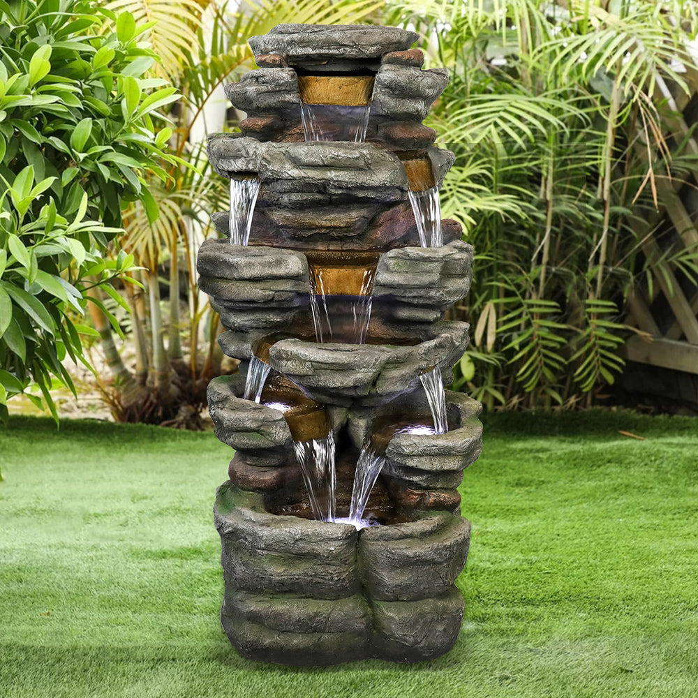 Resin Stepped Shale Outdoor Rock Fountain stands on a lawn amid dense green garden foliage.