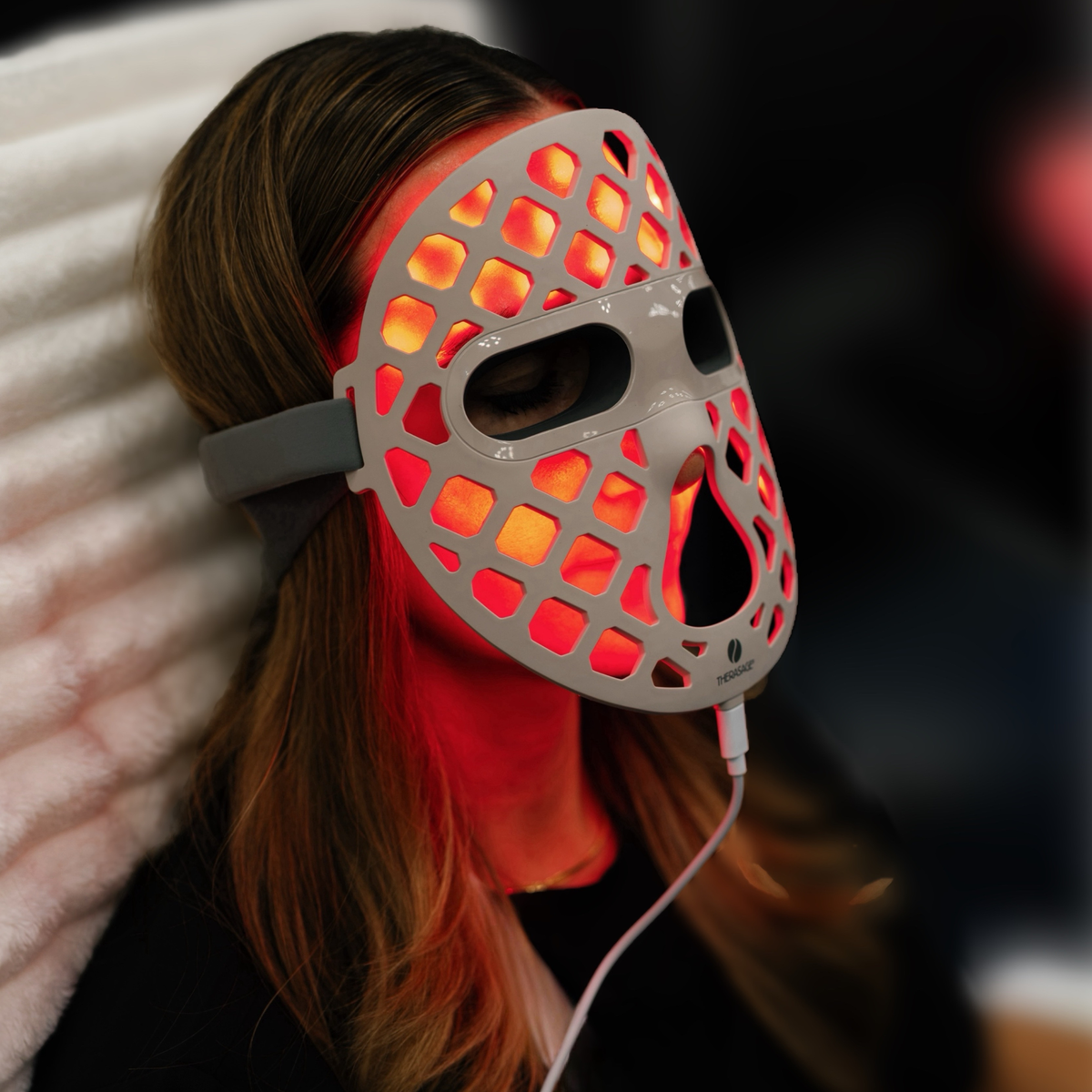 A woman reclines wearing the red-lit TheraGlow Mask2Go+™ LED Light Therapy Face Mask, viewed from the side.