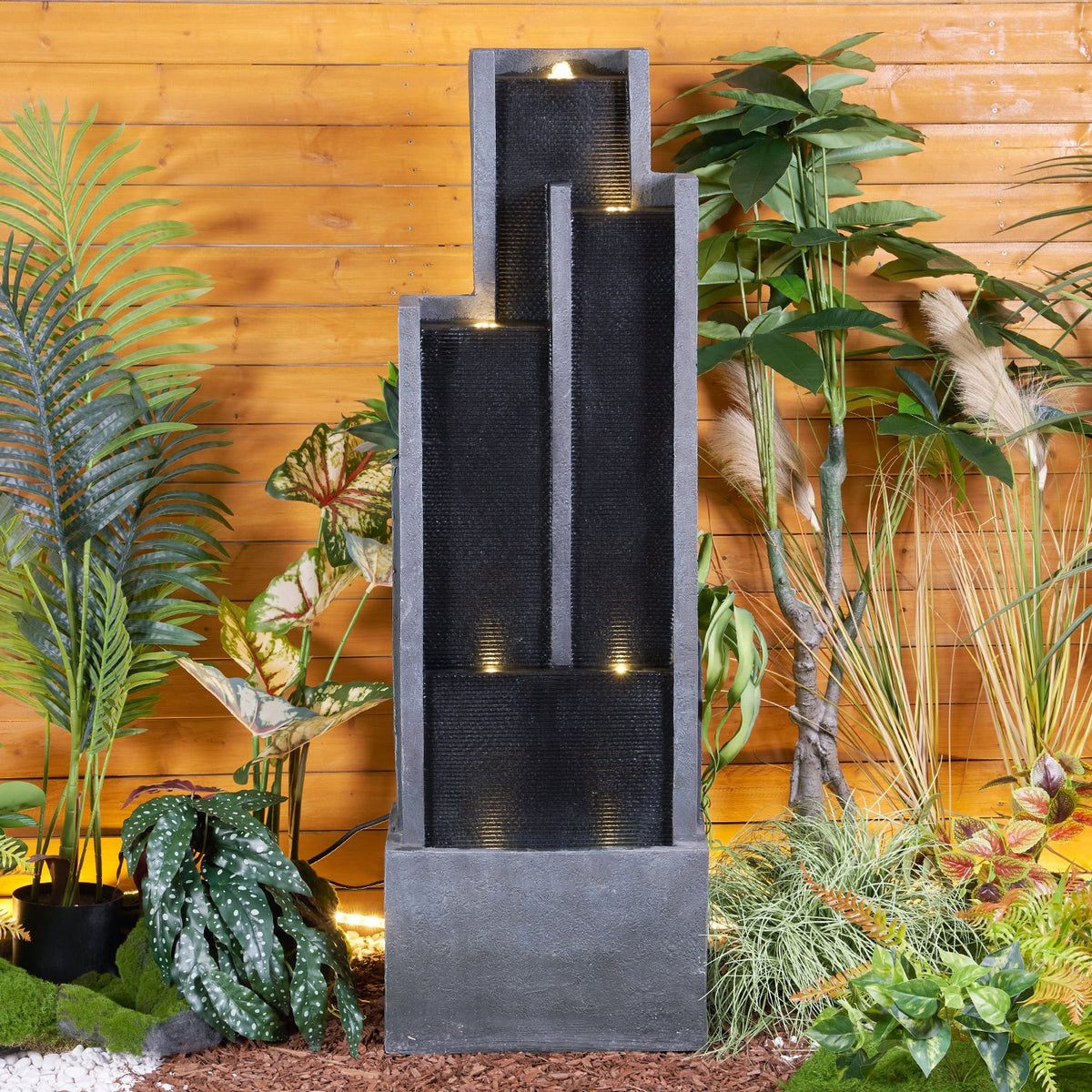 Resin Ribbed Slate Outdoor Water Fountain Quiet 50 in stands among leafy plants before a horizontal wood fence.