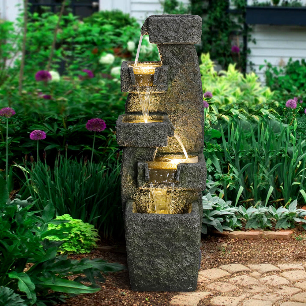 Resin Tiered Outdoor Fountain 5 Tier Modern LED stands in a leafy garden with purple flowers.