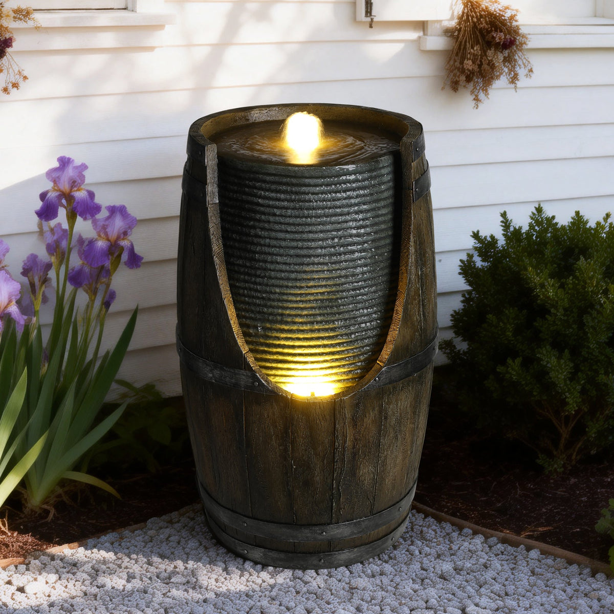 Resin Rustic Barrel Outdoor Water Fountain 24 Inch LED with glowing water, beside purple irises and a shrub.