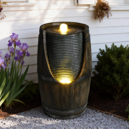 Resin Rustic Barrel Outdoor Water Fountain 24 Inch LED with glowing water, beside purple irises and a shrub.