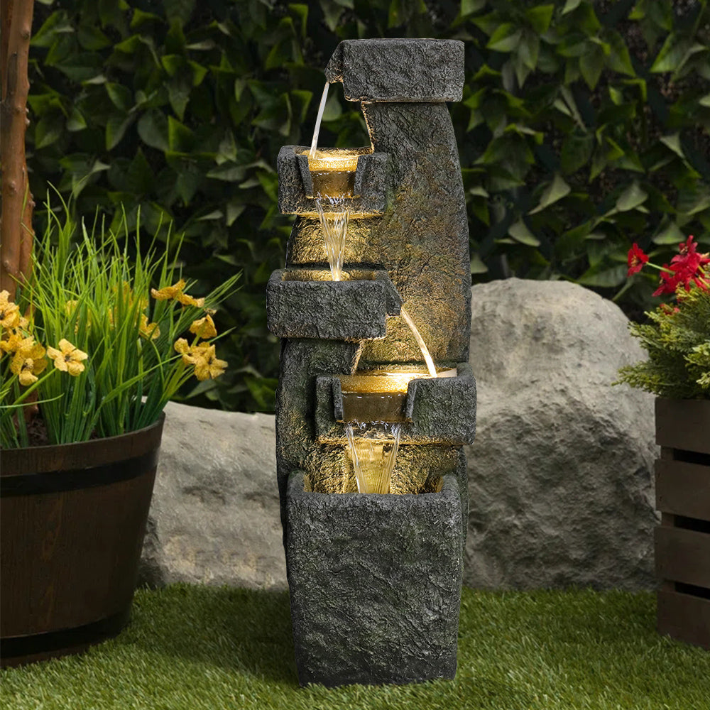 Resin Tiered Outdoor Fountain 5 Tier Modern LED lit among lawn grass, potted flowers, and rocks.