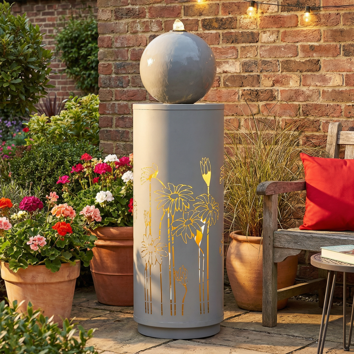 Metal Sphere Water Fountain Outdoor with a glowing floral-patterned column in a brick patio garden beside a chair.